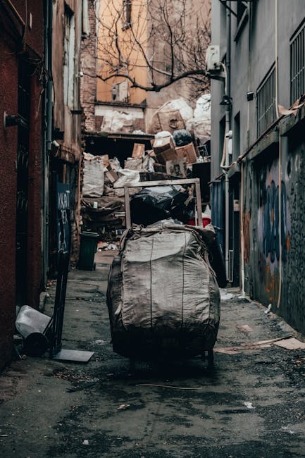 In this outdoor scene, a large collection of mixed household and office waste is piled around three rubbish bins on a paved street. The central bin is a grey, metal container marked for mixed paper an